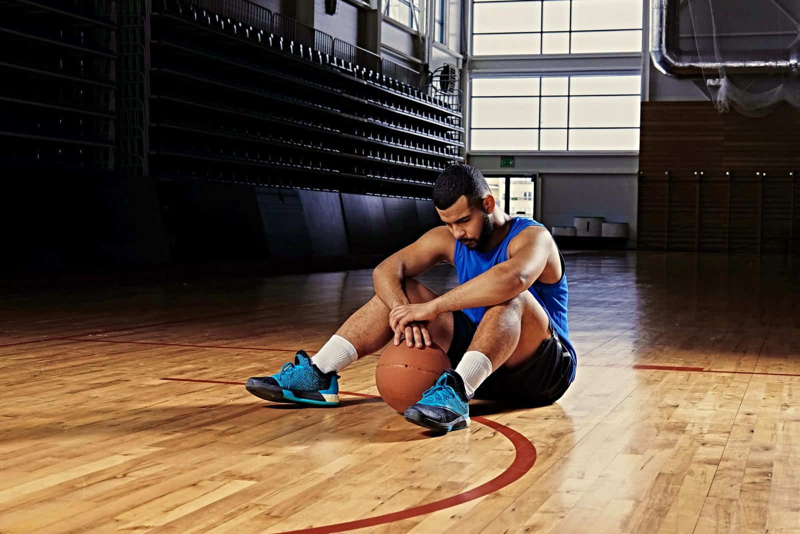 Exhausted basketball player sitting mid-court, recovering from foot pain due to lack of proper shock-absorbing insoles.